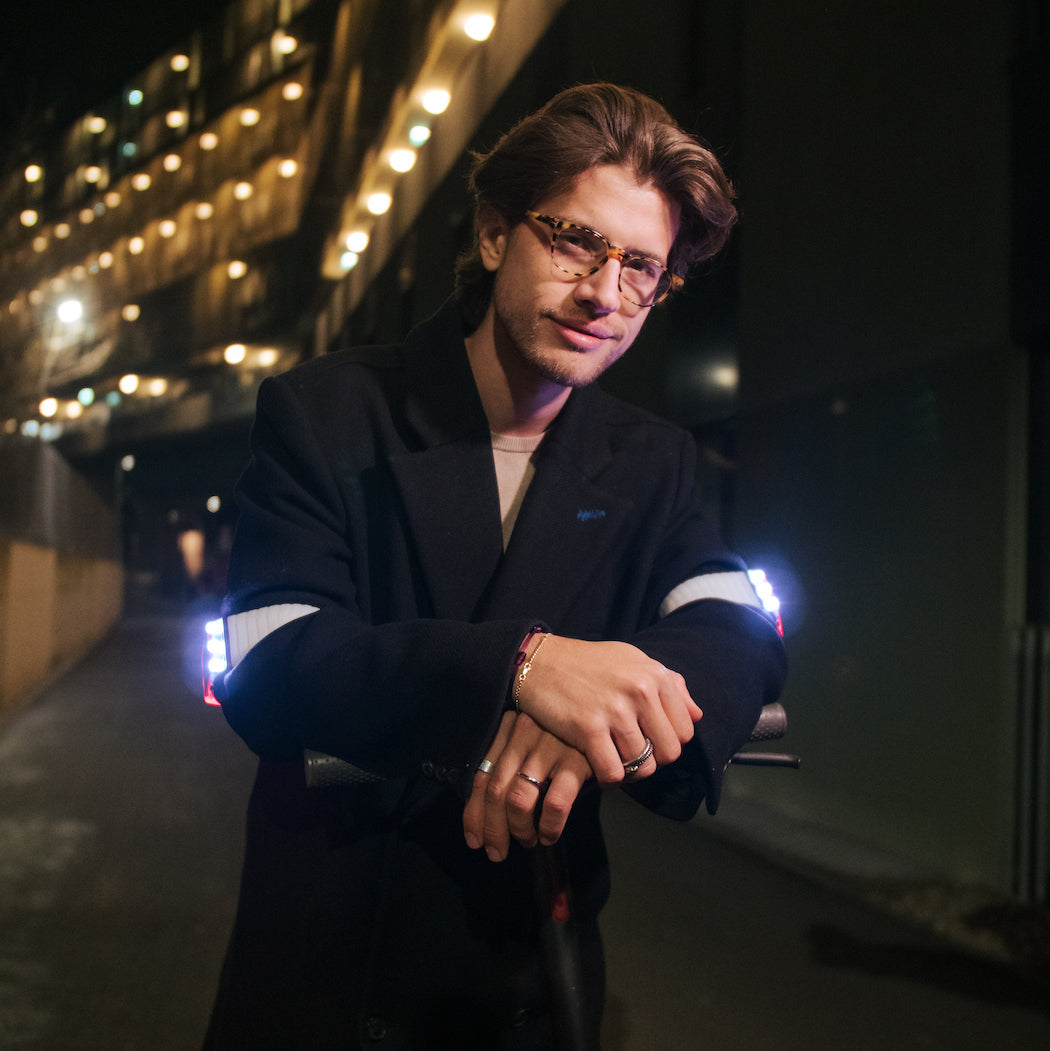 Junger Mann in dunklem Wintermantel, nachts, lehnt lächelnd und mit Flasher Armbändern um die Oberarme, die hell weiß leuchten, auf Lenker eines E-Scooters.