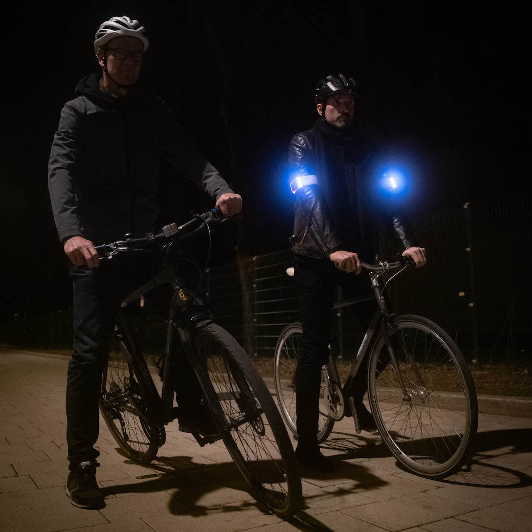 Zwei Radfahrer nachts mit dunkler Kleidung. Radfahrer links ist schwer sichtbar, radfahrer rechts ist von zwei Flasher Armbändern extrem hell beleuchtet, die weiß um seine Oberarme leuchten.