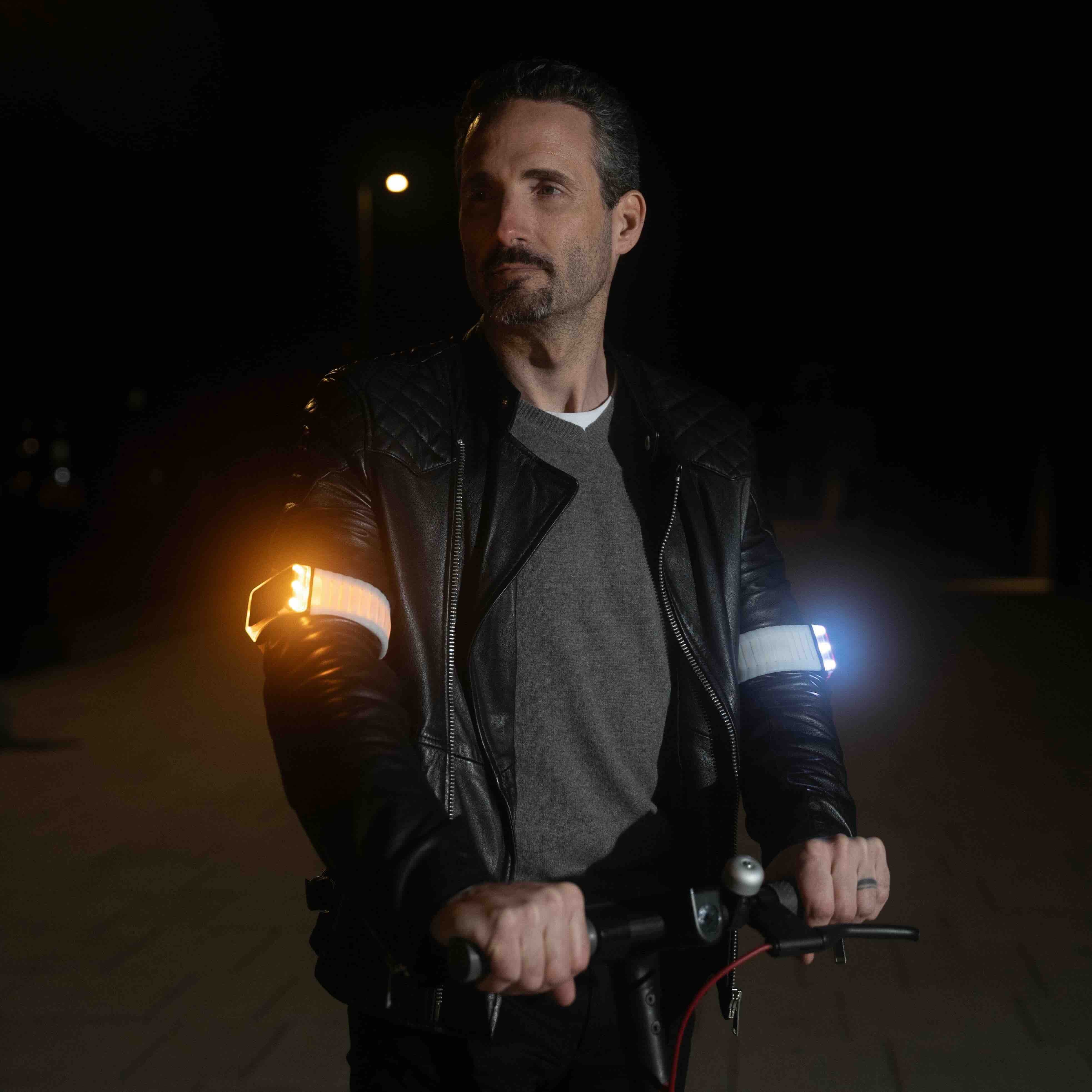 Älterer Herr mit schwarzer Lederjacke nachts steht auf E-Scooter und trägt zwei strahlende Flasher Armbänder, wovon das rechte weiß leuchtet und das linke gelb blinkt.