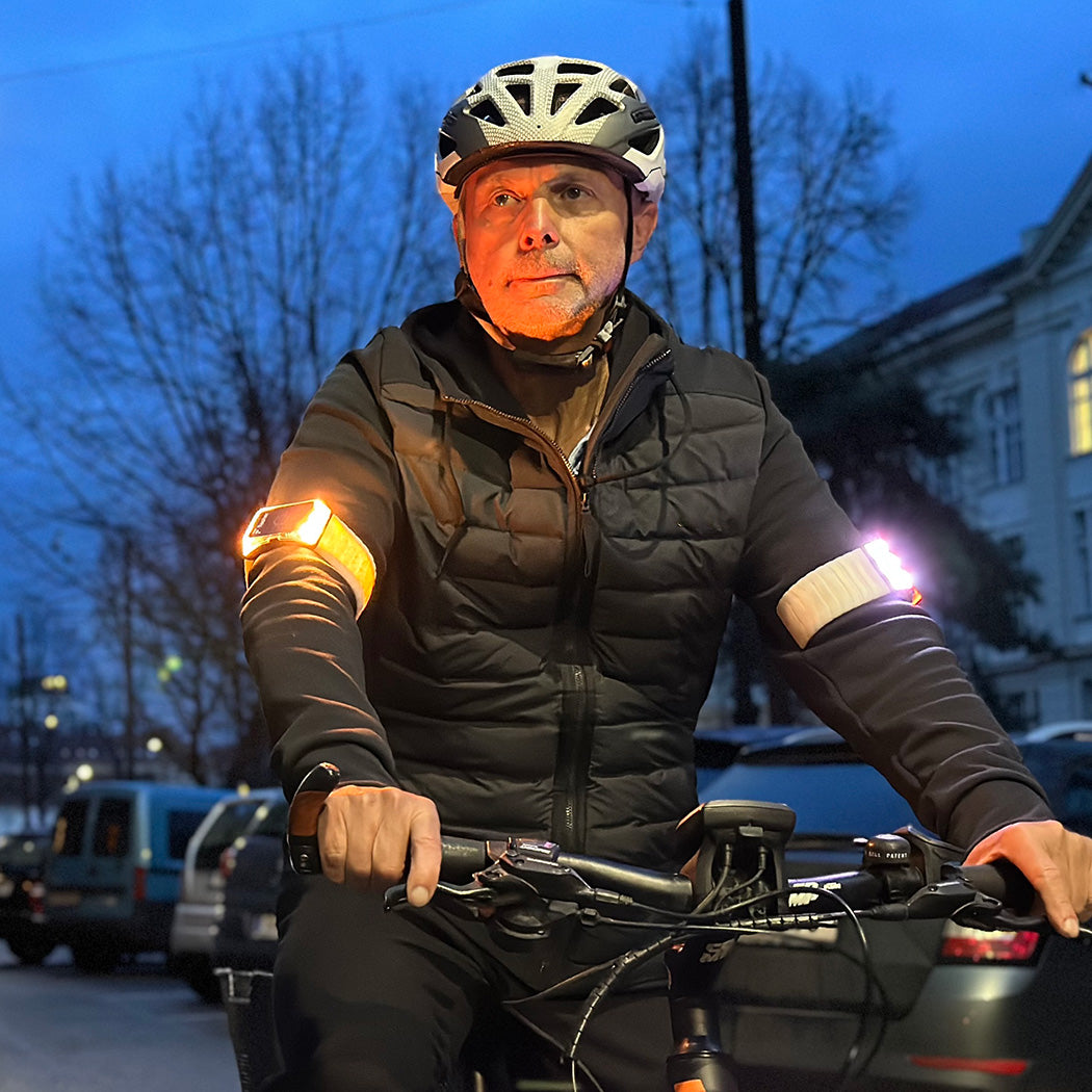 Älterer Radfahrer in der Dämmerung mit dunkler Kleidung, der hell leuchtende Flasher Armbänder trägt. Rechts leuchtet es weiß und rot, links blinkt es gelb.