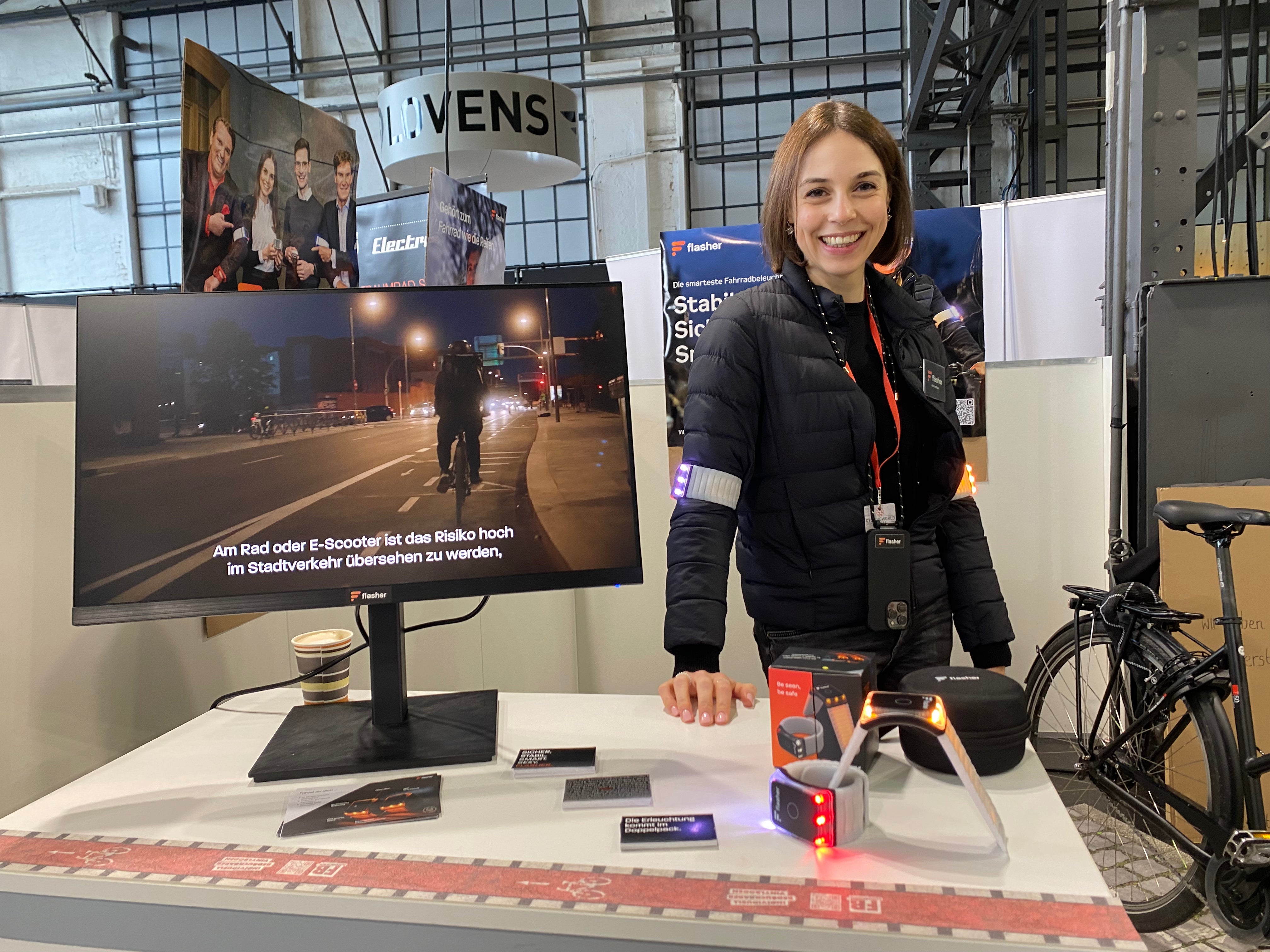 Ines auf unserem Messestand auf der Cyclingworld Düsseldorf 2024, mit Flasher Armbändern um die Oberarme und auf der weißen Theke vor sich, neben einem Monitor auf dem ein Erklärvideo läuft und vor einem Posten mit dem Text "Stabil. Sichtbar. Smart."