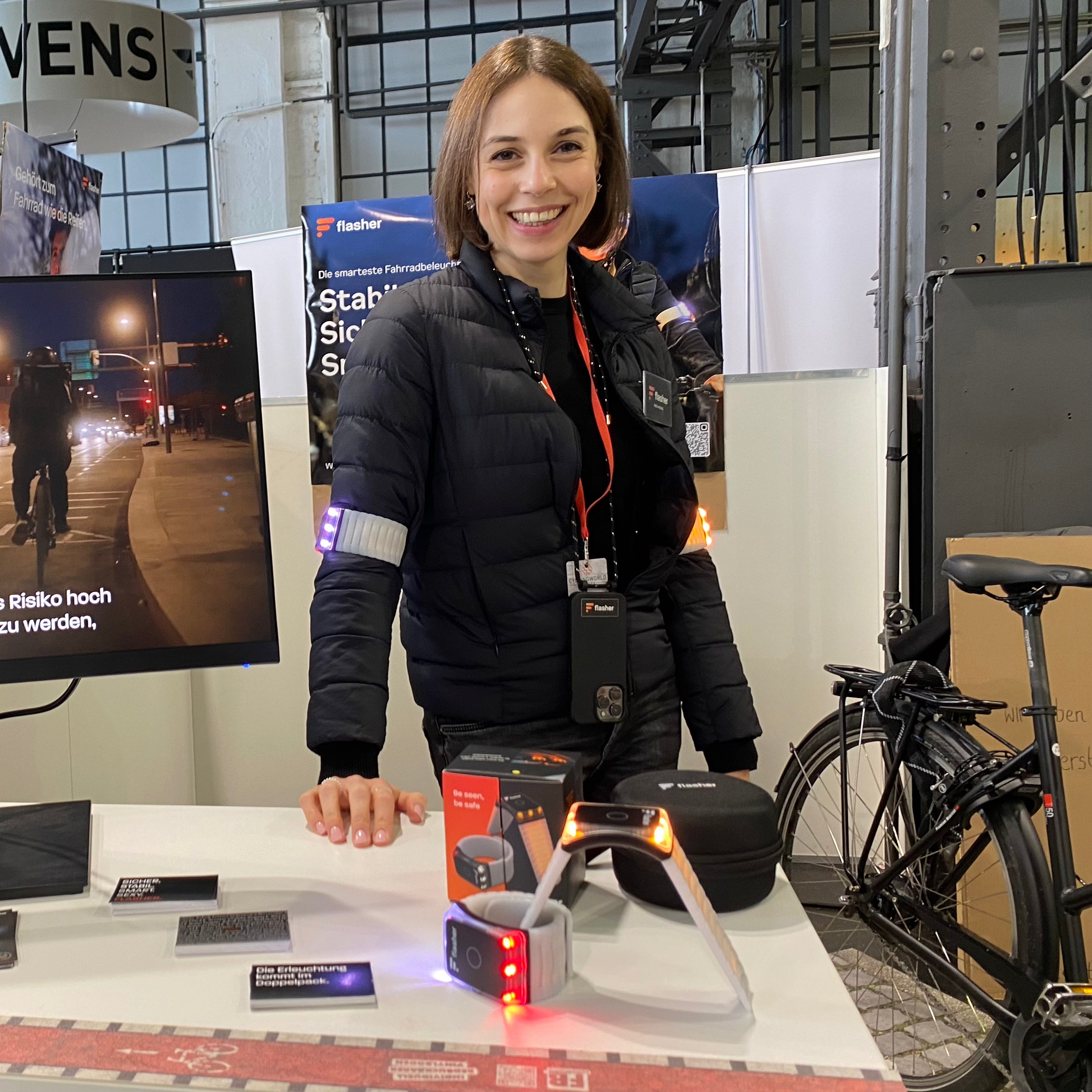 Ines auf unserem Messestand auf der Cyclingworld Düsseldorf 2024, mit Flasher Armbändern um die Oberarme und auf der weißen Theke vor sich, neben einem Monitor auf dem ein Erklärvideo läuft und vor einem Posten mit dem Text "Stabil. Sichtbar. Smart."