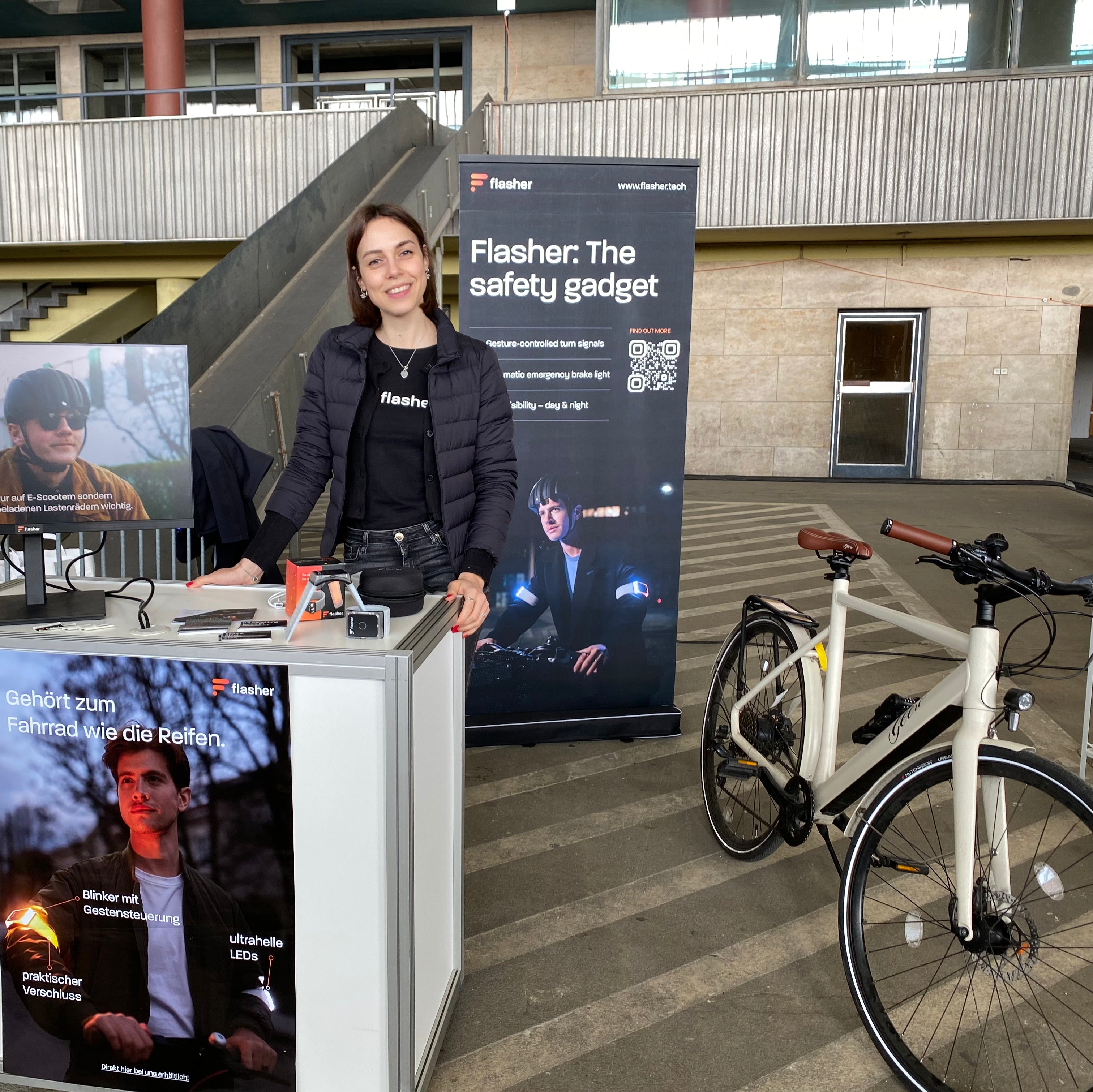 Ines auf unserem VELOBerlin-Stand, vor einer weißen Theke mit einem Flasher Set, einem Monitor auf dem ein Erklärungsvideo läuft und neben einem Fahrrad von Geero.