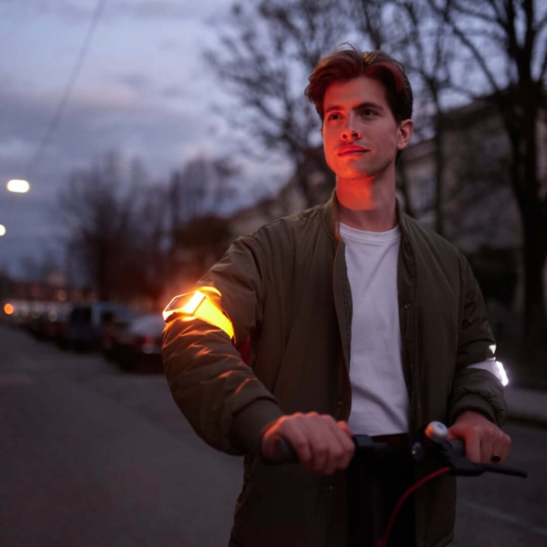 Junger Mann in Dämmerung auf E-Scooter mit zwei Flasher Armbändern, das rechte leuchtet weiß und das linke blinkt strahlend gelb.