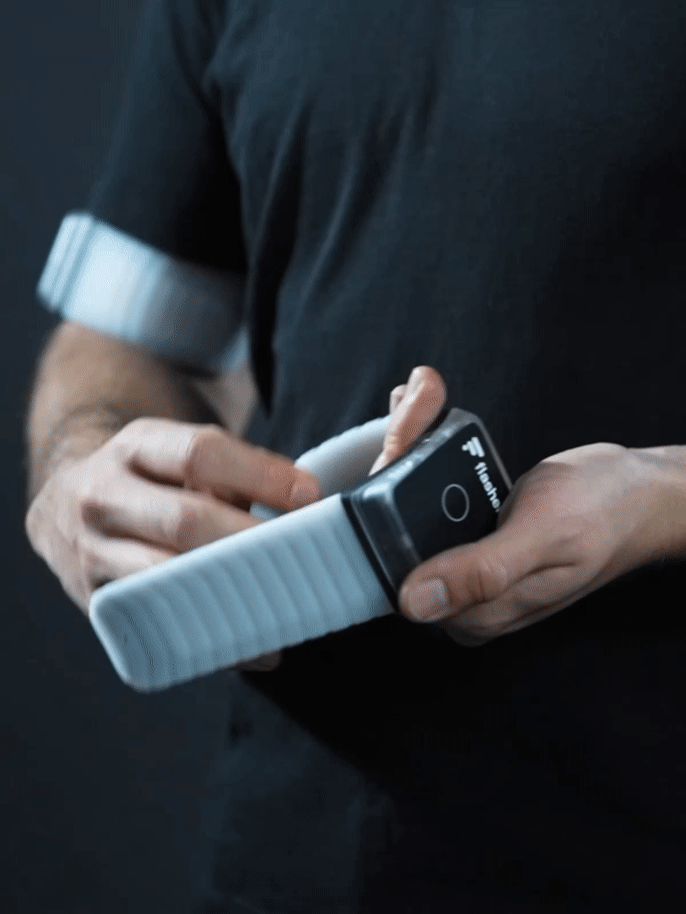 Person klappt die Flügel eines Flasher Armbands auf, legt das Armband knapp ober dem Ellbogen an den Oberarm an und schnappt beide Flügel mit einem kurzen Druck zu.