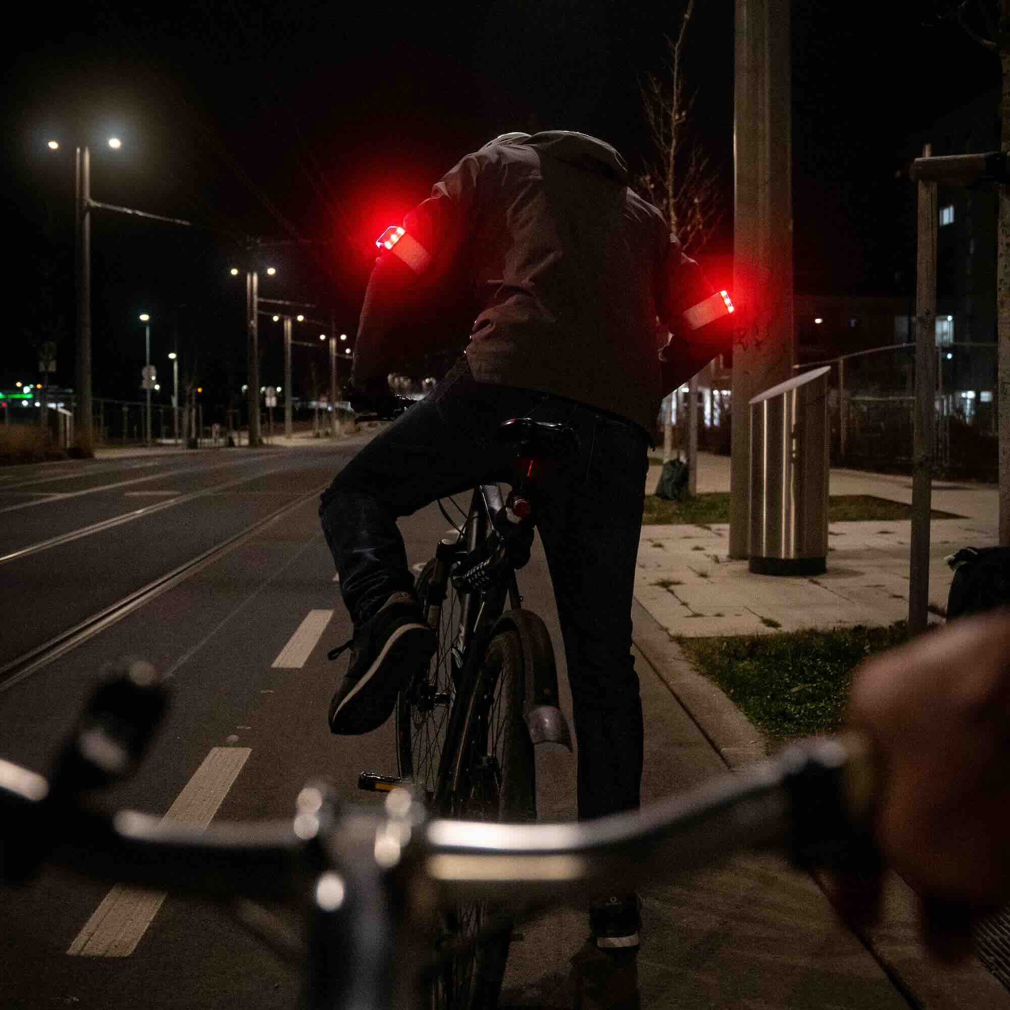 Radfahrer im Straßenverkehr von hinten, nachts, mit zwei Flasher Armbändern auf den Oberarmen. Das Bremslicht der Armbänder leuchtet auf beiden Seiten hell rot.