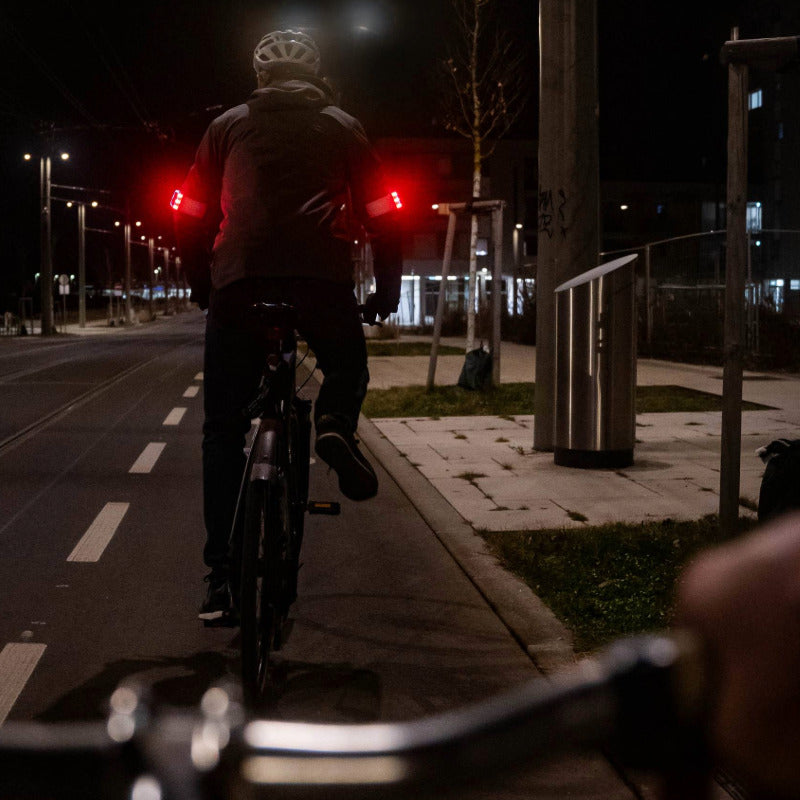 Radfahrer im Straßenverkehr von hinten, nachts, mit zwei Flasher Armbändern auf den Oberarmen, die auf beiden Seiten hell rot leuchten.